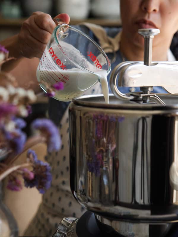 A person pouring vegan buttermilk from a measuring cup into a shiny metal stand mixer. Purple flowers are blurred in the foreground, and the person is wearing a white apron with a blue shirt.